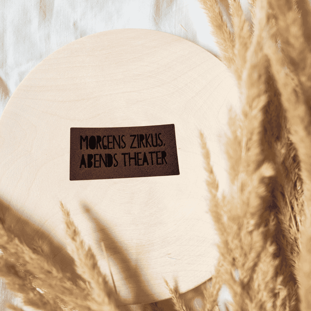 Rechteckiges dunkelbraunes Kunstleder-Label mit dem Spruch „Morgen Zirkus, Abends Theater“, ca. 4,7 x 2,3 cm, veganes Leder-Patch für humorvolle Nähprojekte, laser-graviert bei Schnuckidu