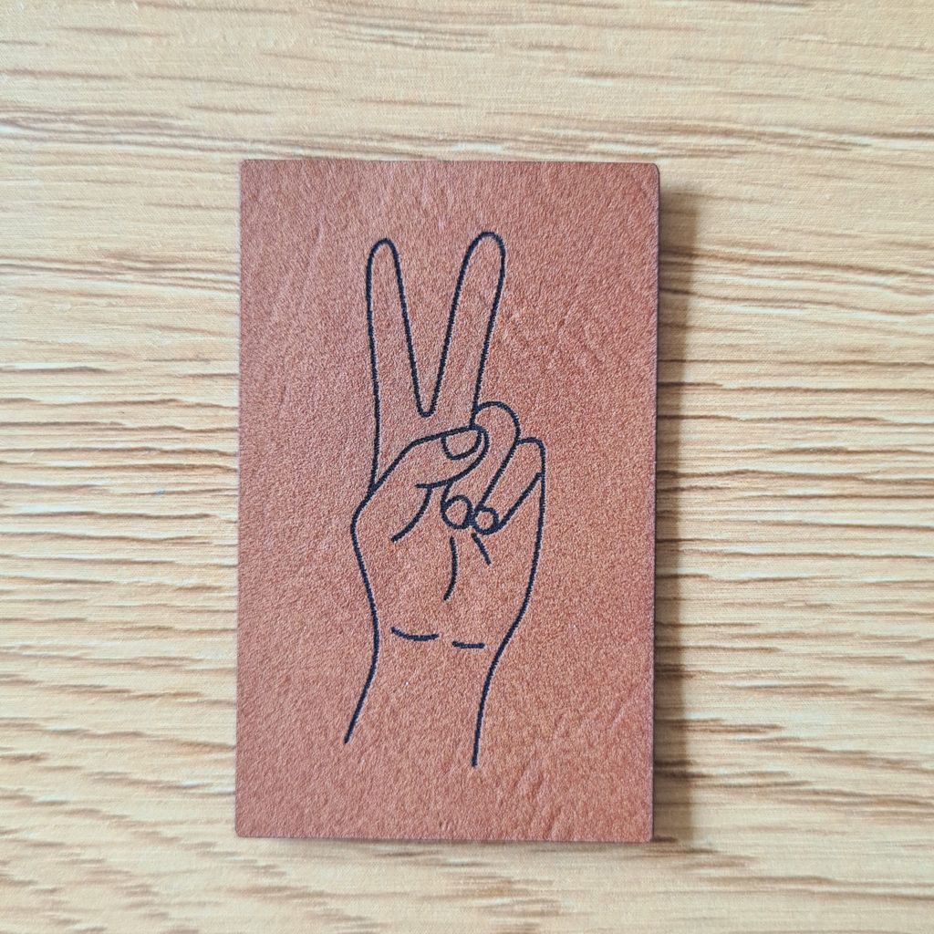 Hochformatiges braunes Kunstleder-Label mit einem fein lasergravierten Peace-Handzeichen, veganer Aufnäher für Hoodies und Taschen, laser-gravierte Eigenproduktion von Schnuckidu.