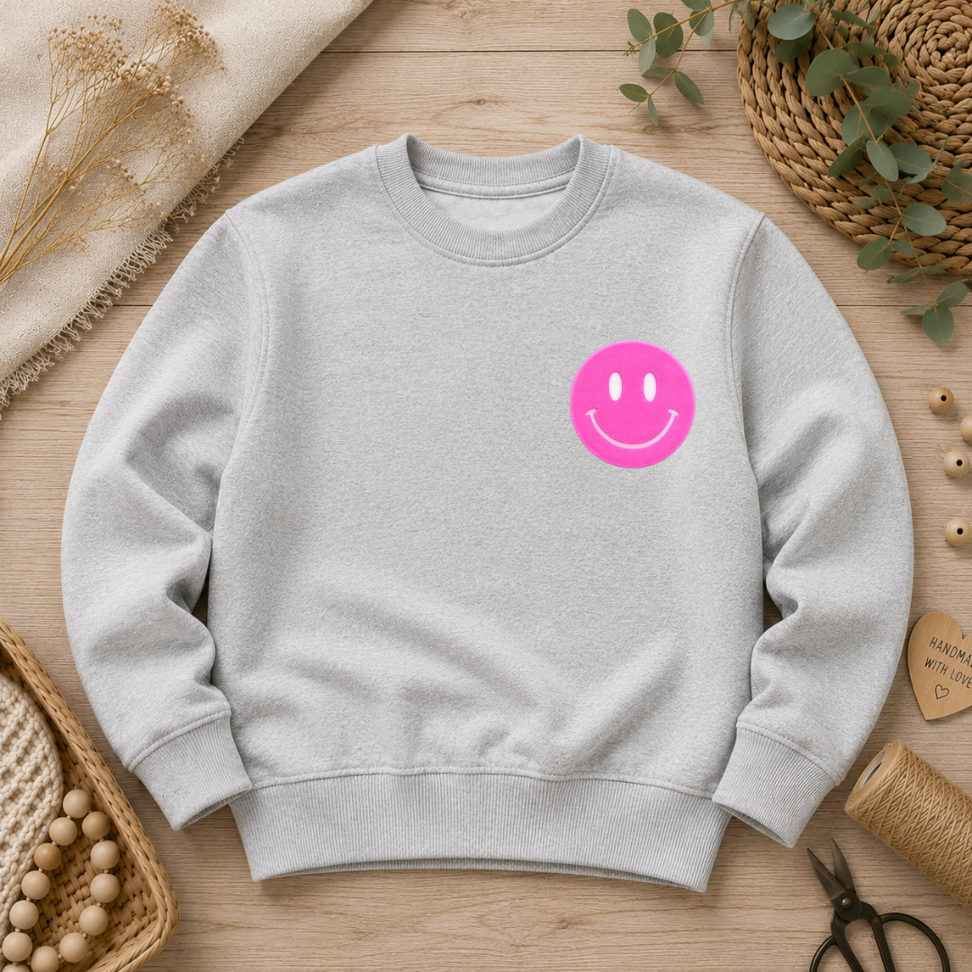 Samtiges, kreisrund ausgefülltes Flock-Patch in Form eines gut gelaunten Smileys in Neon-Pink, aufgebügelt auf der Brust eines hellgrauen Sweatshirts.