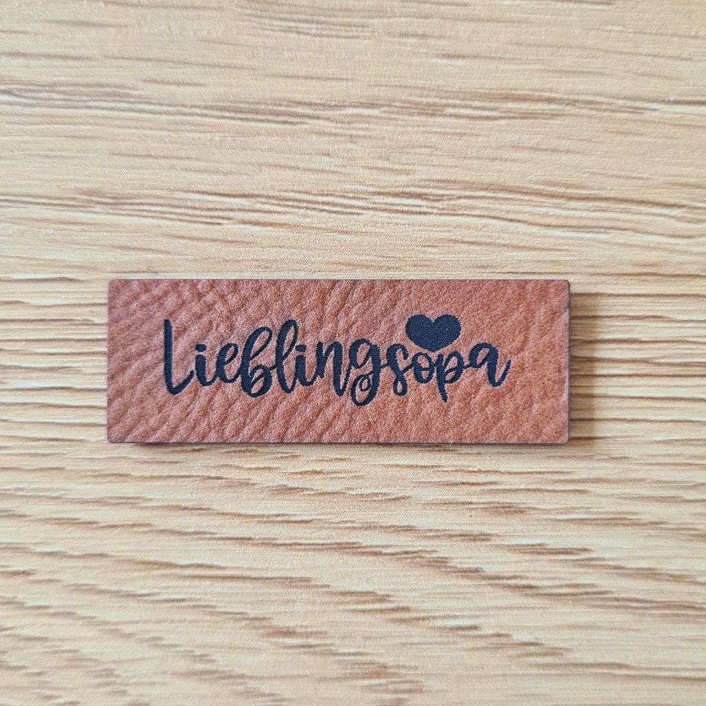 Rechteckiges braunes Kunstleder-Label mit dem fein gravierten geschwungenen Schriftzug „Lieblingsopa“ und einem kleinen ausgefüllten Herz-Detail, veganes Nählabel für handgemachte Geschenke, laser-graviert bei Schnuckidu.
