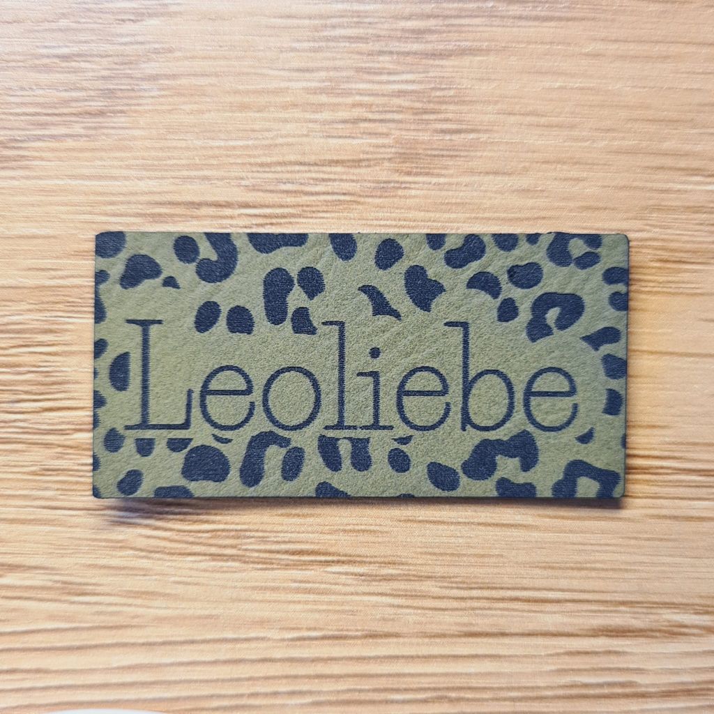 Rechteckiges olivegrünes Kunstleder-Label mit dem Schriftzug „Leoliebe“ und dezenten Leoparden-Splotches, 5 x 2,5 cm, veganes Nählabel für moderne Kleidung, laser-graviert bei Schnuckidu.