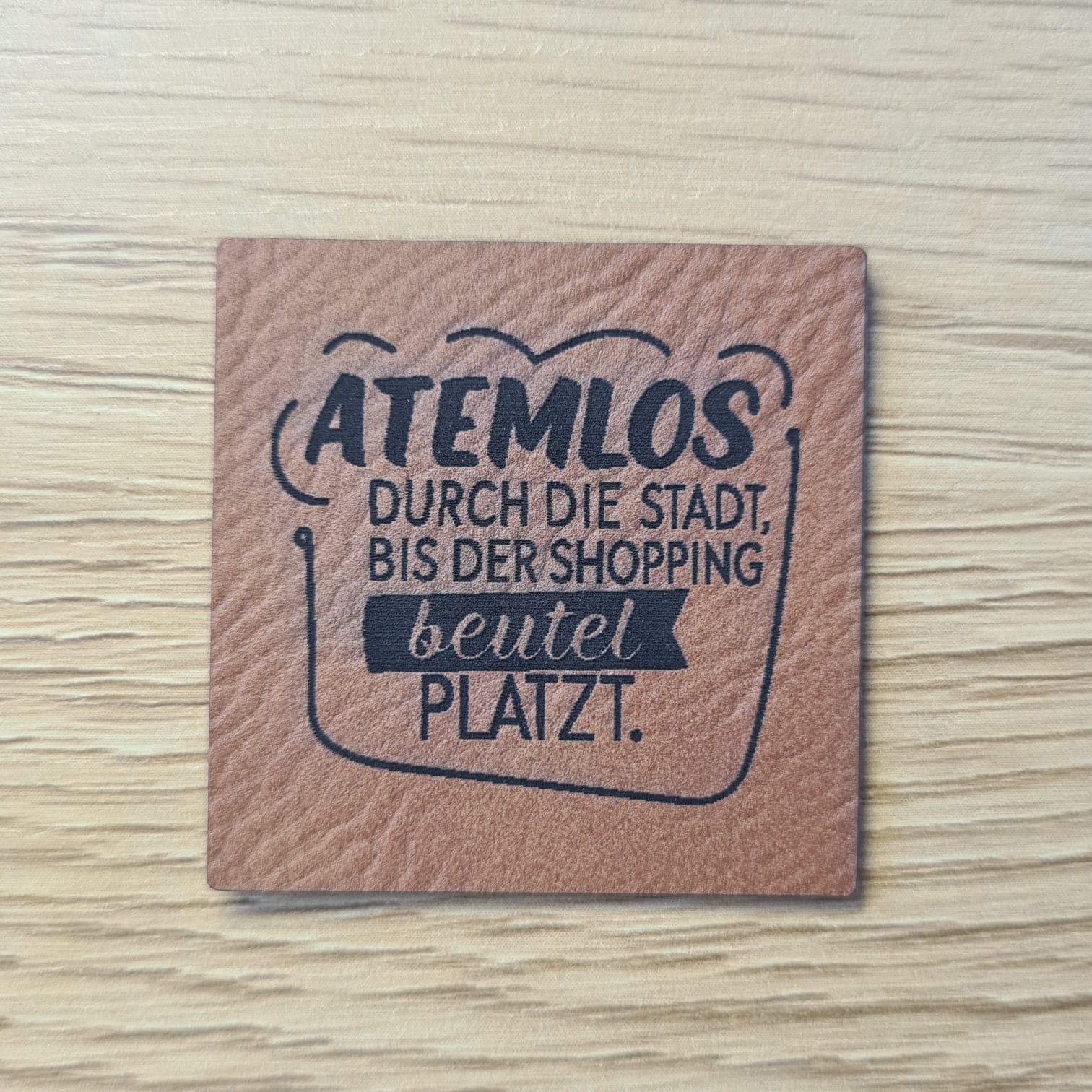 Schnuckidu Kunstleder Nählabel in Braun quadratisch mit Henkeltaschen-Rahmen und dem Spruch Atemlos durch die Stadt, bis der Shoppingbeutel platzt.