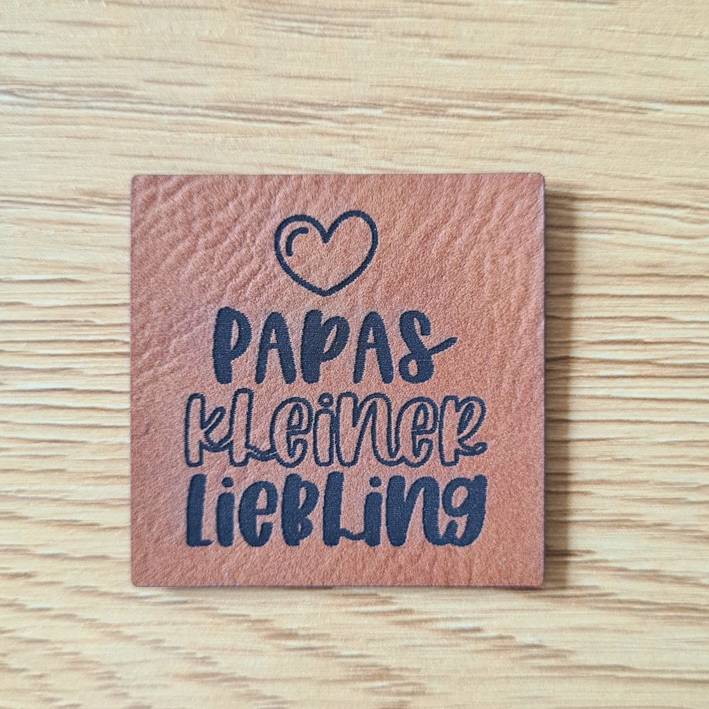 Quadratisches braunes Kunstleder-Label mit dem fein gravierten Schriftzug „Papas kleiner Liebling“, veganer Aufnäher für Baby- und Kinderkleidung, laser-gravierte Eigenproduktion von Schnuckidu.