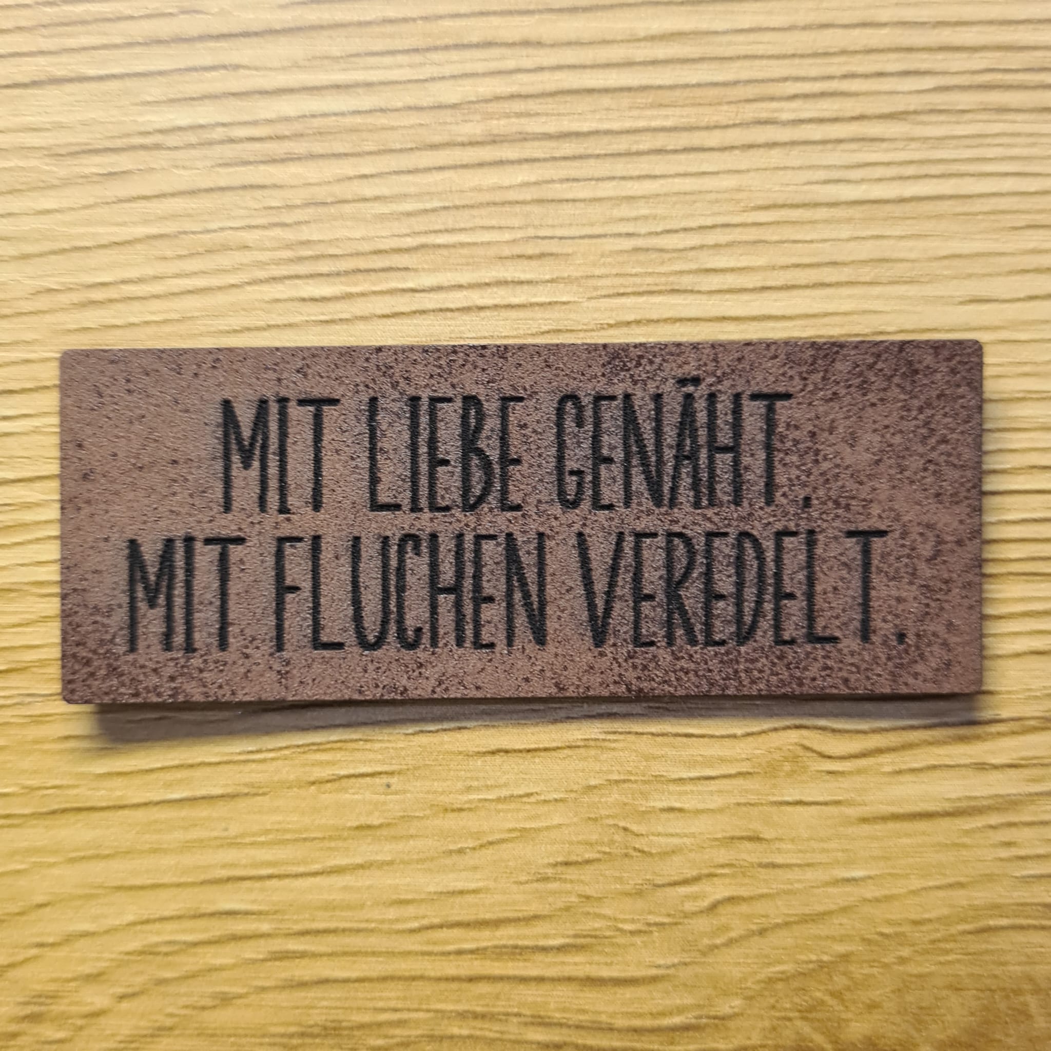 Großes dunkelbraunes Kunstleder-Label mit dem Spruch „Mit Liebe genäht, mit Fluchen veredelt“, ca. 5,8 x 2,2 cm, veganes Leder-Patch für humorvolle Nähprojekte, laser-graviert bei Schnuckidu