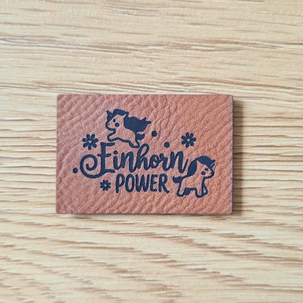 Rechteckiges braunes Kunstleder-Label mit dem fein gravierten Schriftzug „Einhorn POWER“, zwei niedlichen Einhörnern und kleinen Sternen, veganer Aufnäher für Kinderkleidung, laser-graviert bei Schnuckidu