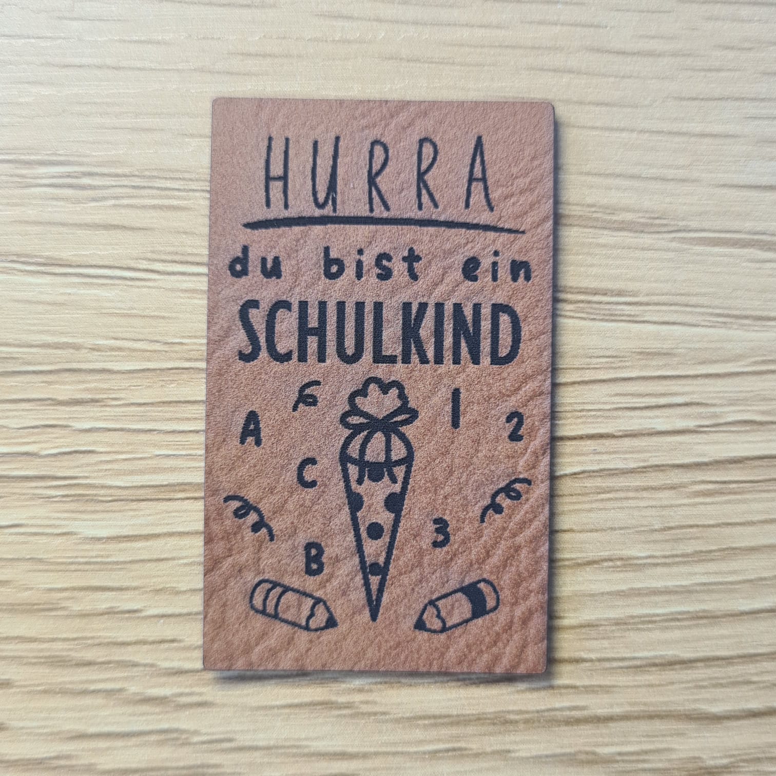 Schnuckidu Kunstleder Nählabel in Braun Hochformat mit Schriftzug Hurra du bist ein Schulkind, Schultüte, Stiften, Buchstaben und Zahlen.