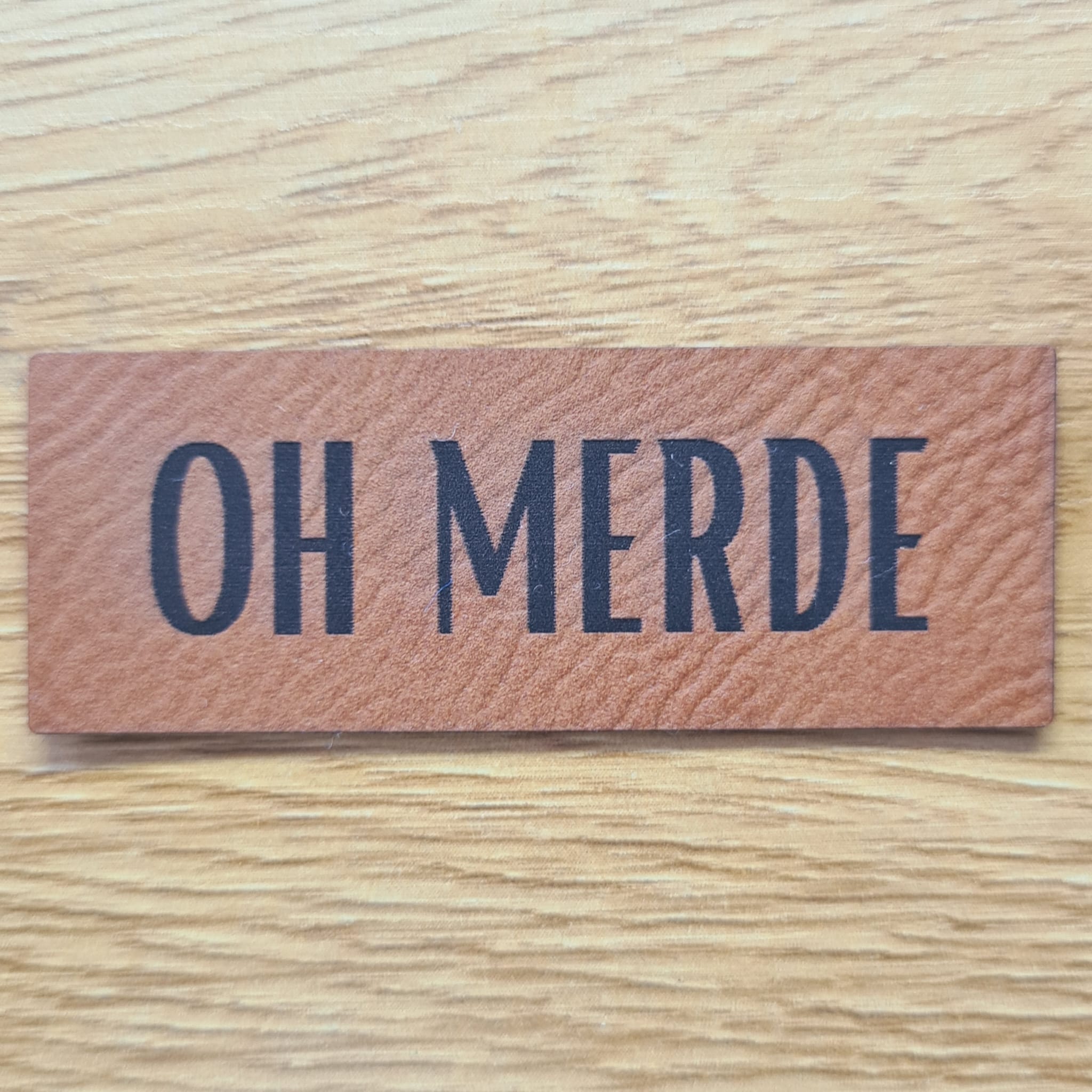 Rechteckiges braunes Kunstleder-Label mit dem gravierten Spruch „OH MERDE“, 6,0 x 2,2 cm, veganes Nählabel mit Humor für Kleidung und Taschen, laser-graviert bei Schnuckidu.