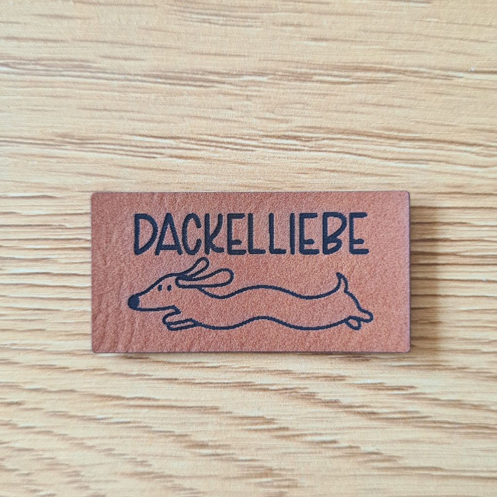 Rechteckiges braunes Kunstleder-Label mit dem fein gravierten Schriftzug „DACKELLIEBE“ und einer gezeichneten, fliegenden Dackelsilhouette darunter, veganer Aufnäher für Hundezubehör und Kleidung, laser-graviert bei Schnuckidu.