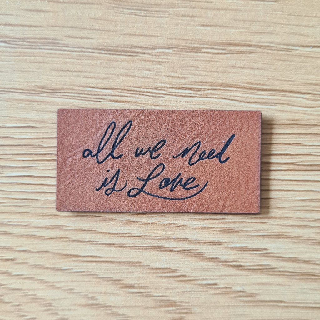 Rechteckiges braunes Kunstleder-Label mit dem fein gravierten Spruch „all we need is Love“, veganer Aufnäher für Kleidung und Accessoires, laser-gravierte Eigenproduktion von Schnuckidu.