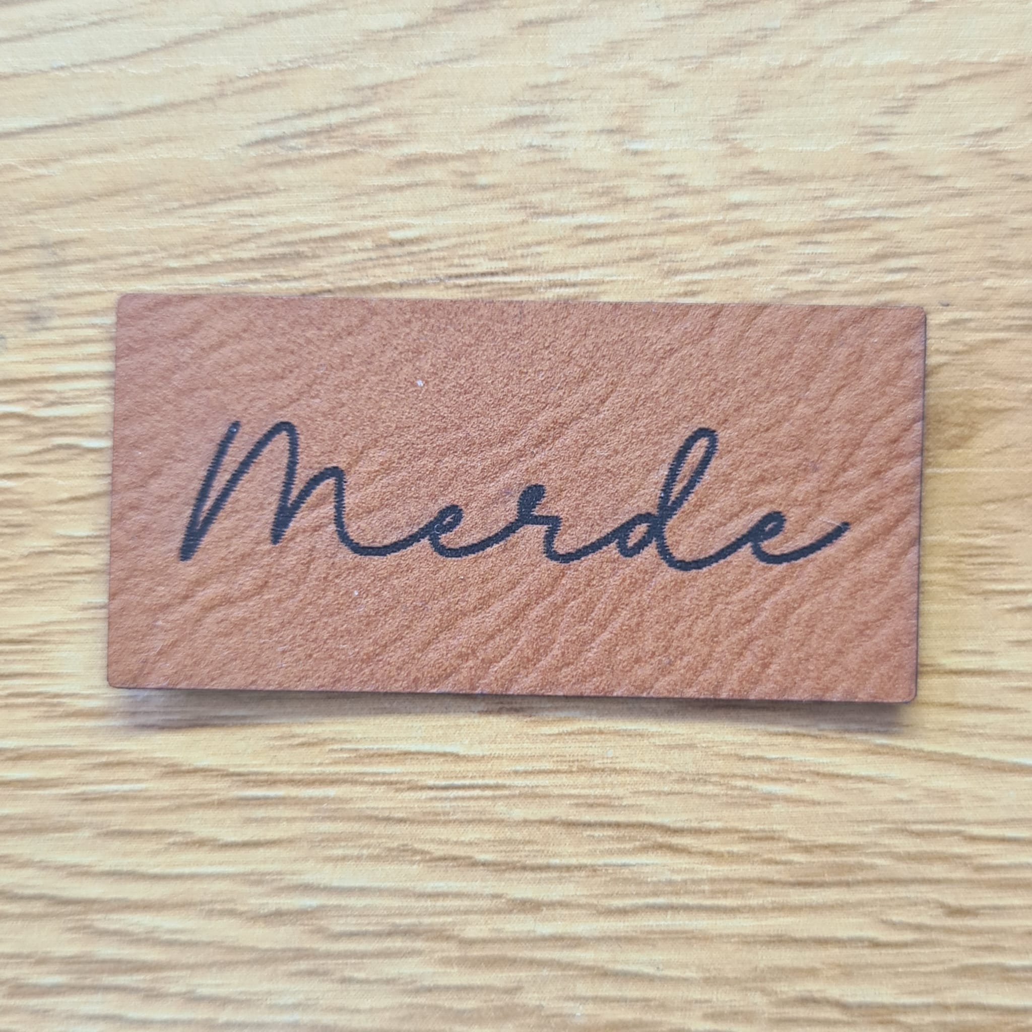 Rechteckiges braunes Kunstleder-Label mit dem in eleganter Schreibschrift gravierten Wort „merde“, 4,5 x 2,2 cm, veganes Nählabel mit Humor für Kleidung und Taschen, laser-graviert bei Schnuckidu.