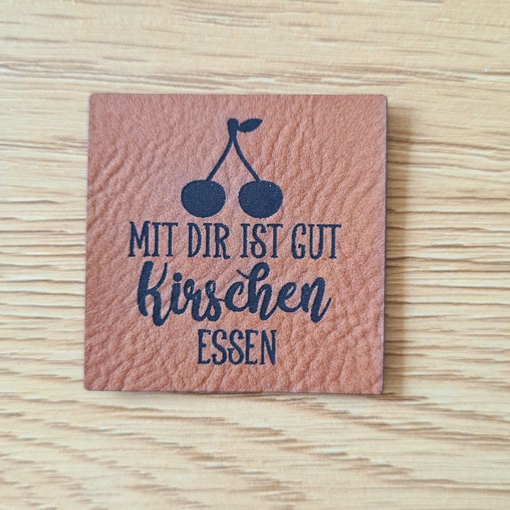 Quadratisches braunes Kunstleder-Label mit dem fein gravierten Spruch „Mit dir ist gut Kirschen essen“, veganer Aufnäher für Kleider und sommerliche Outfits, laser-gravierte Eigenproduktion von Schnuckidu