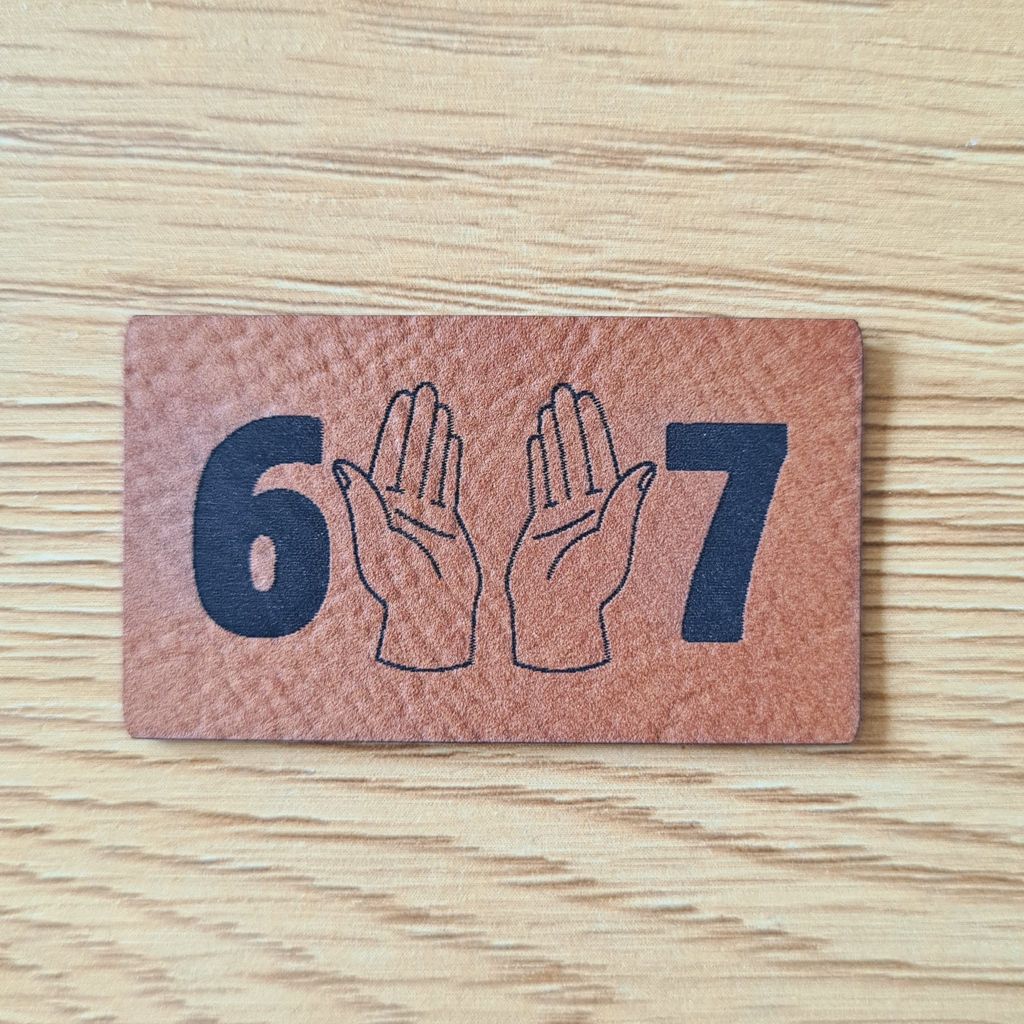Rechteckiges braunes Kunstleder-Label im Querformat mit der markanten Zahl 67 und zwei zentral gravierten, geöffneten Händen, veganer Aufnäher für Streetwear und Hoodies, laser-gravierte Eigenproduktion von Schnuckidu.