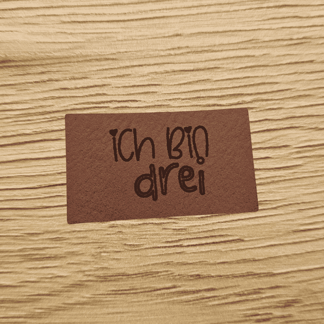 Rechteckiges braunes Kunstleder-Label mit dem gravierten Kinderschriftzug „Ich bin drei“ und kleinen Herz-Details, bezugnehmend auf das Produktbild ichbindrei.png, veganes Nählabel für Kinderkleidung, laser-graviert bei Schnuckidu.