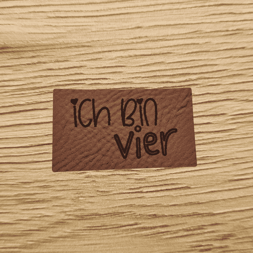 Rechteckiges braunes Kunstleder-Label mit dem gravierten Kinderschriftzug „Ich bin vier“ und kleinen Herz-Details, bezugnehmend auf das Produktbild ichbinvier.png, veganes Nählabel für Kinderkleidung, laser-graviert bei Schnuckidu.