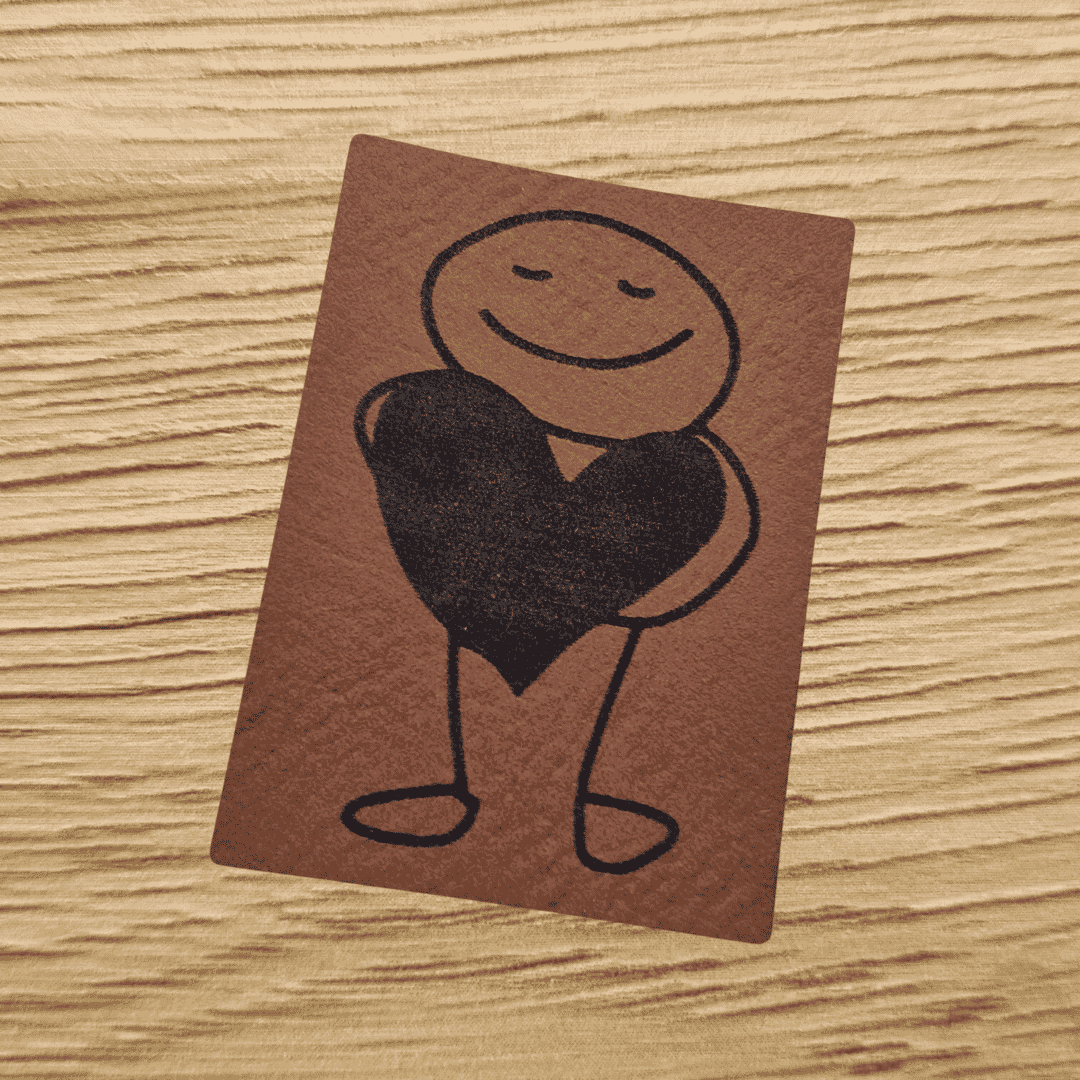 Rechteckiges braunes Kunstleder-Label mit einer gravierten Strichfigur, die ein großes Herz hält, 3,1 x 5 cm, veganes Nählabel für Kleidung und Taschen, laser-graviert bei Schnuckidu.