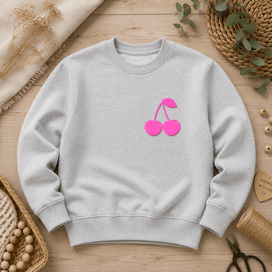 Samtiges, dreidimensionales Flock-Patch in Form von zwei Kirschen mit Blatt in Neon-Pink aufgebügelt auf der Brust eines grauen Sweatshirts.