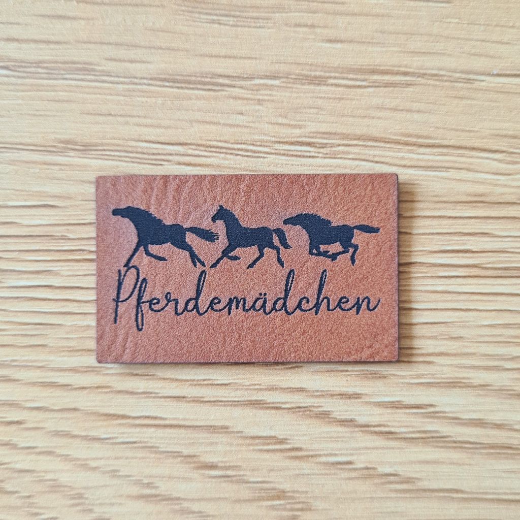 Rechteckiges braunes Kunstleder-Label mit dem fein gravierten Schriftzug „Pferdemädchen“ und drei galoppierenden Pferdesilhouetten darüber, veganer Aufnäher für Reitkleidung, Jacken und Accessoires, laser-graviert bei Schnuckidu.