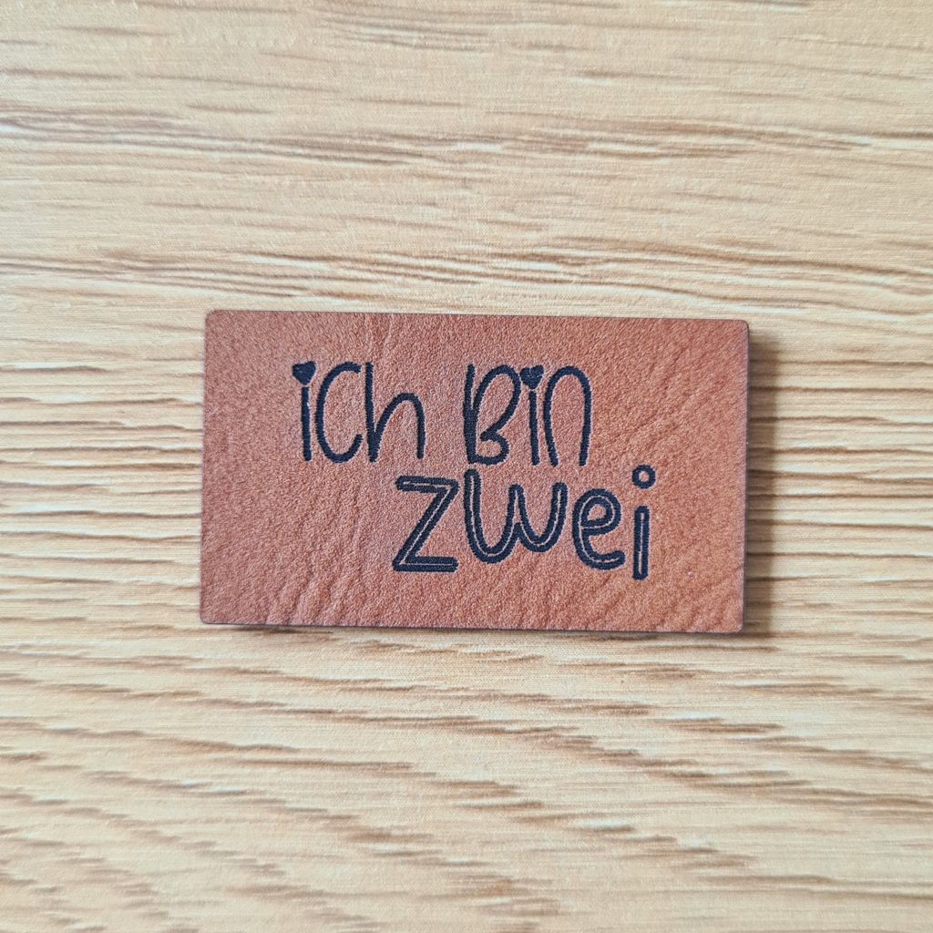 Rechteckiges braunes Kunstleder-Label mit dem gravierten Kinderschriftzug „Ich bin zwei“ und kleinen Herz-Details, bezugnehmend auf das Produktbild ichbinzwei.png, veganes Nählabel für Kinderkleidung, laser-graviert bei Schnuckidu.