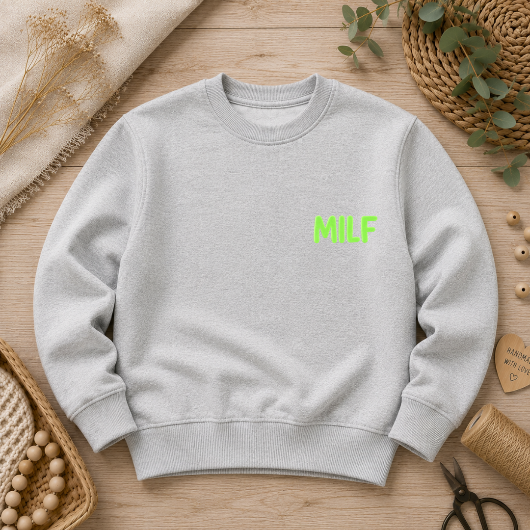 Samtiges Flock-Patch mit dem prägnanten Schriftzug "MILF" in leuchtendem Neon-Grün, aufgebügelt auf der Brust eines hellgrauen Sweatshirts.