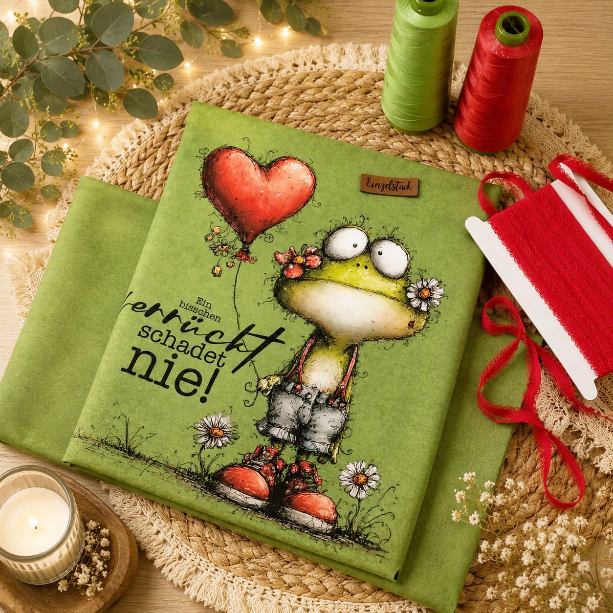 Grünes Stoffpanel mit gezeichnetem Frosch, der ein rotes Herz-Luftballon hält, umgeben von Gänseblümchen und dem Text 'Ein bisschen verrückt schadet nie!'
