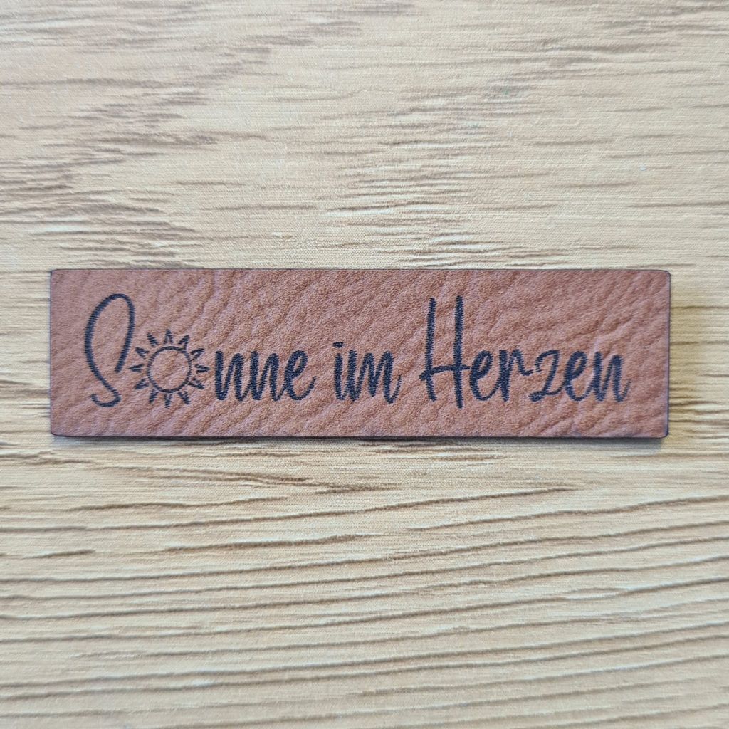 Schmales, rechteckiges braunes Kunstleder-Label mit dem Schriftzug „Sonne im Herzen“, 5,4 x 1,5 cm, veganes Nählabel für zuversichtliche Wohlfühlkleidung, laser-graviert bei Schnuckidu.
