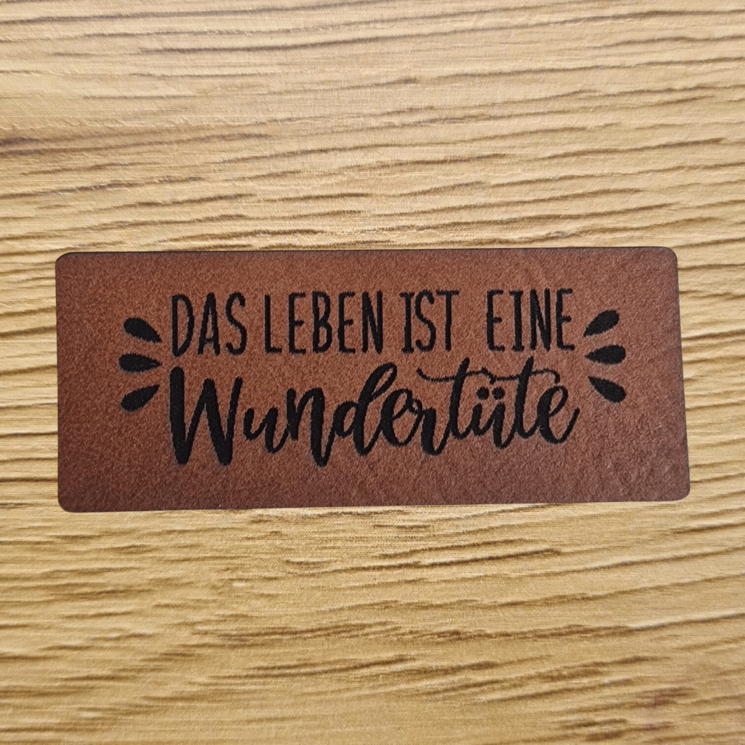 Rechteckiges braunes Kunstleder-Label mit dem gravierten Spruch „Das Leben ist eine Wundertüte“, 5,0 x 2,0 cm, veganes Nählabel für Kleidung und Taschen, laser-graviert bei Schnuckidu.