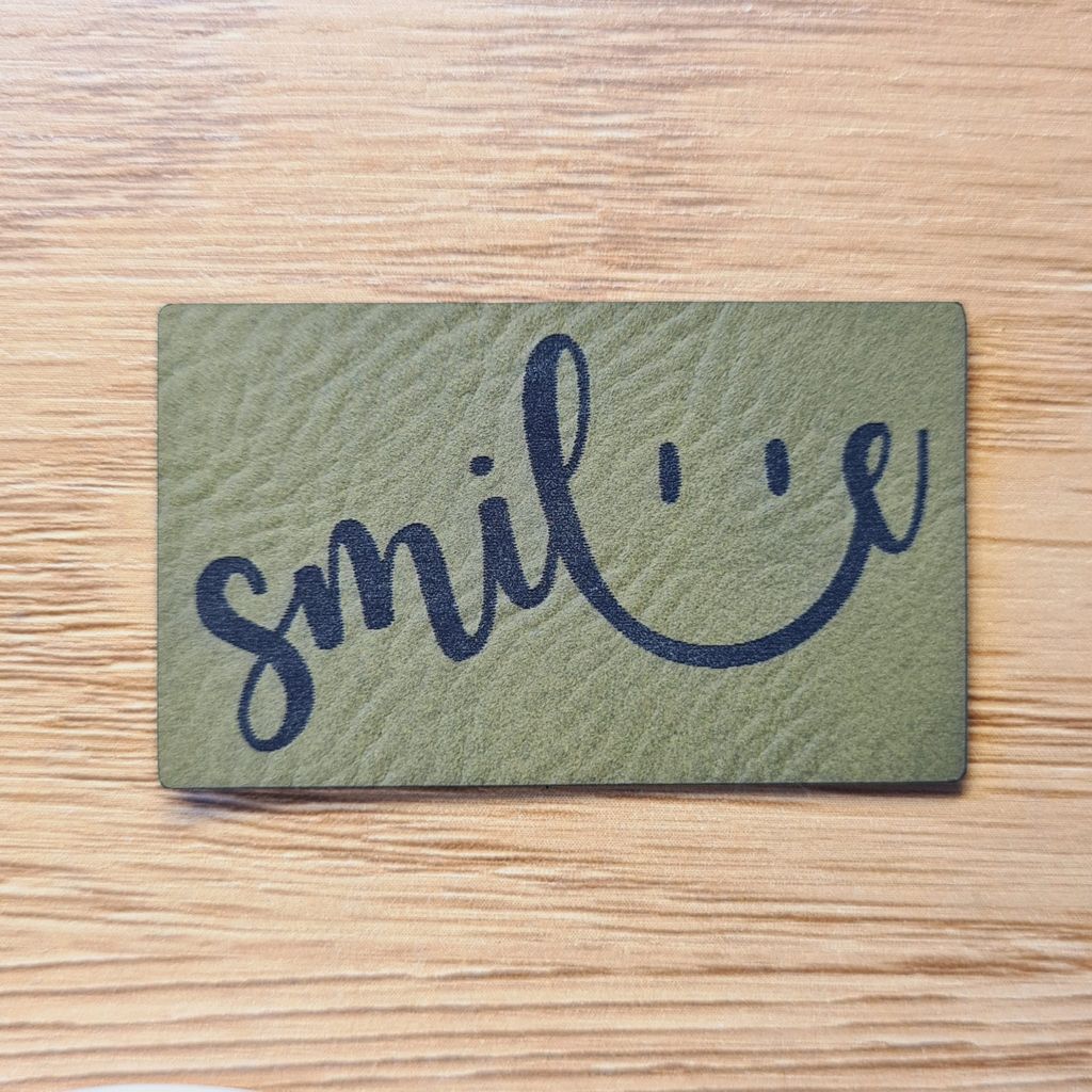 Rechteckiges olivegrünes Kunstleder-Label mit dem geschwungenen Schriftzug „smile“, dessen Bögen ein Smiley-Gesicht bilden, 5 x 3,1 cm, veganes Nählabel für fröhliche Kleidung, laser-graviert bei Schnuckidu.