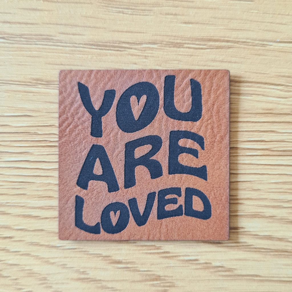 Quadratisches braunes Kunstleder-Label mit dem fein gravierten, modernen Schriftzug „You are loved“, veganer Aufnäher für Kleidung und Accessoires, laser-gravierte Eigenproduktion von Schnuckidu.